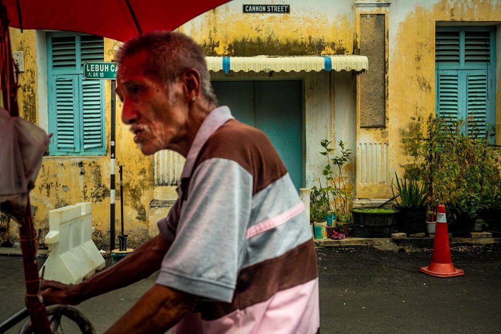 Older man in a striped shirt in the foreground in front of a yellow wall with teal shutters and plants in Penang