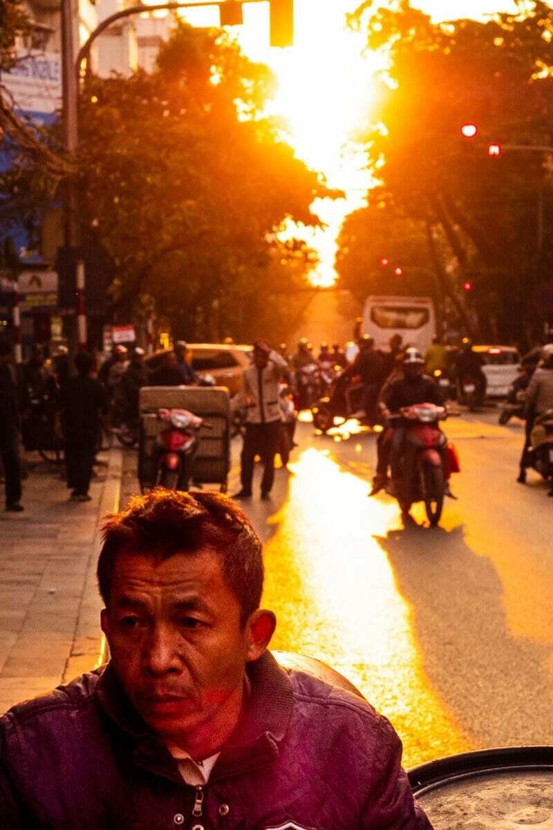 Man's face close in the foreground at the bottom of the frame with a sunset street full of motorbikes glowing behind him in Hanoi