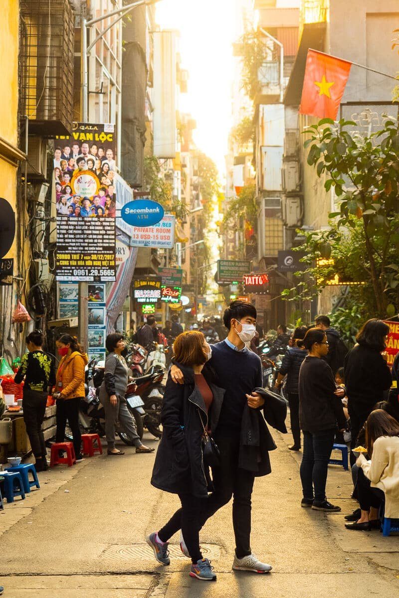 Busy sunlit alley in Hanoi with people walking toward the camera under colorful signs