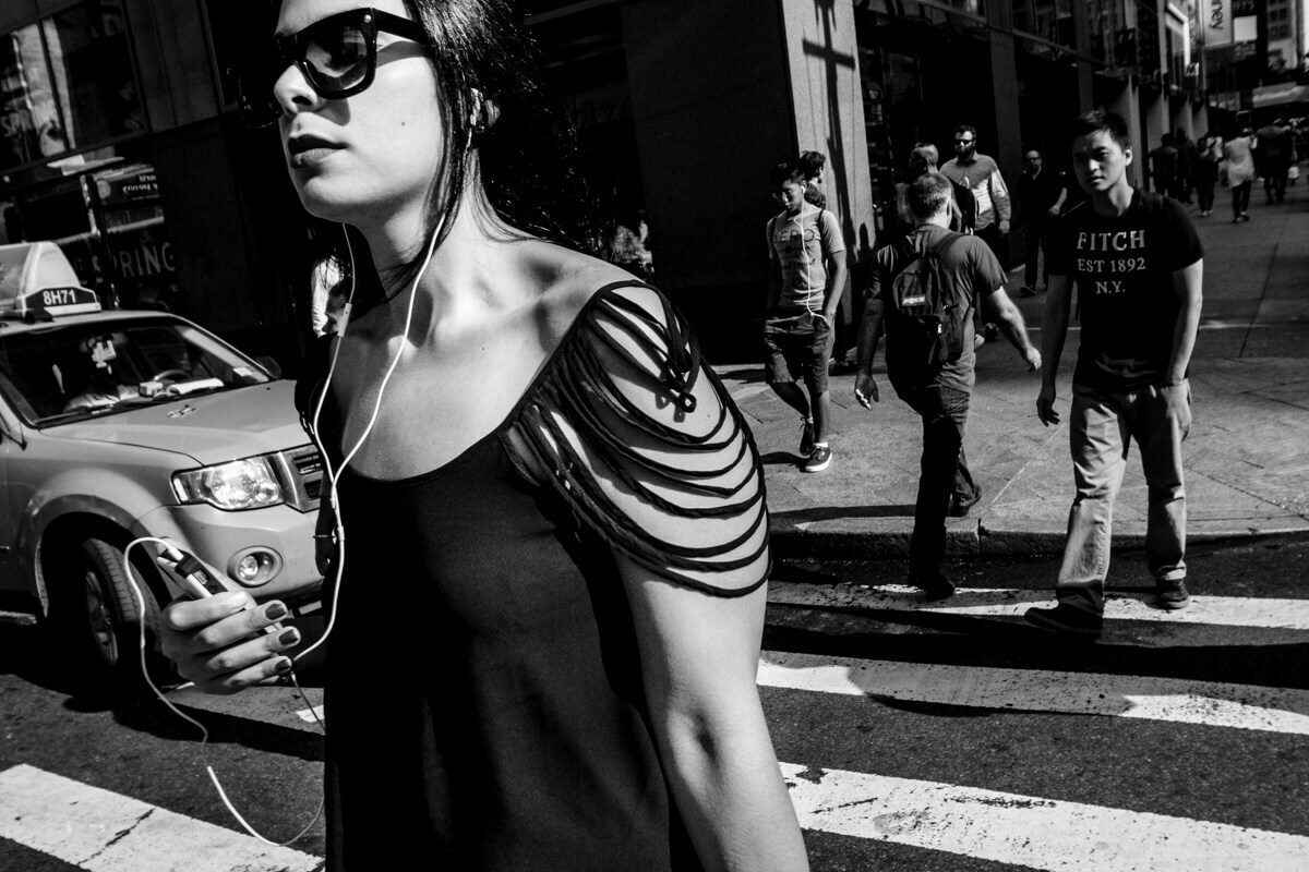 Black and white street photo of a young woman in sunglasses with earphones crossing a busy street beside a taxi