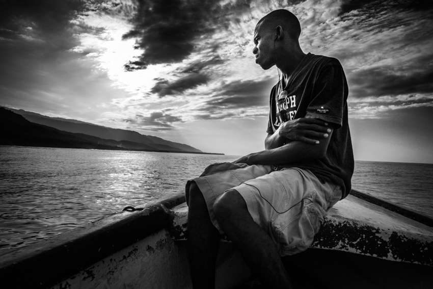 Black and white photo of a young man sitting on the edge of a boat by the sea under a dramatic sky