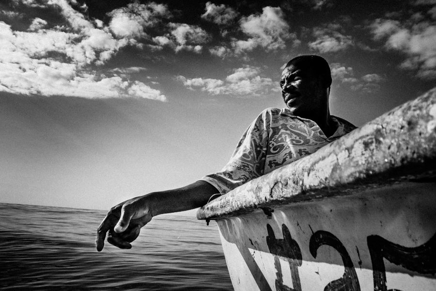 Black and white photo of a man leaning over the side of a painted boat with the sea behind him