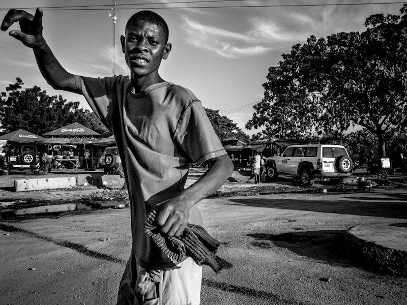 Black and white street photo in Haiti of a young man waving his arm, close to the camera, with a market and cars behind him