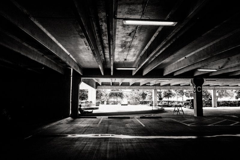 Black and white photo of a dark parking garage with a low concrete ceiling, framing a bright opening onto trees and a street