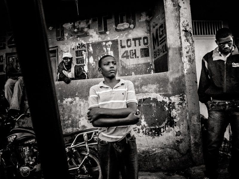 High contrast black and white street photo of a young man with crossed arms standing in front of a weathered wall in Haiti, a man framed in a doorway to the left