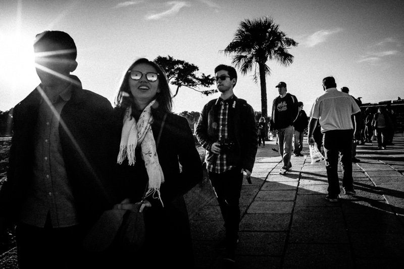 Black and white street photo of people walking toward the camera on a wide sidewalk with the sun flaring behind them and palm trees in the background