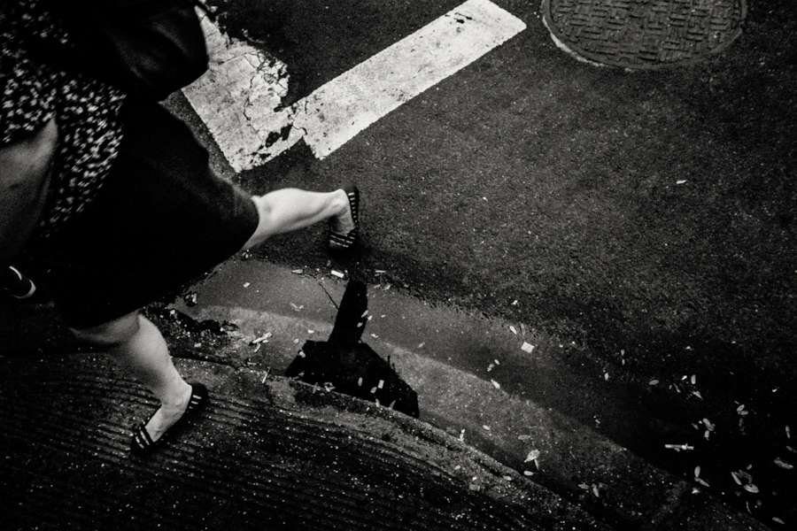 Black and white street photo from above of a woman's legs stepping over a puddle next to a white road marking