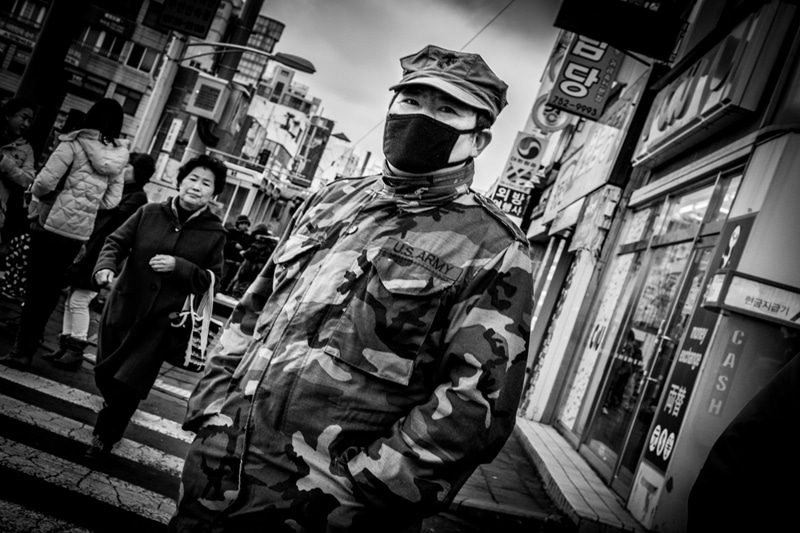 Black and white photo of a man in a camouflage jacket and face mask walking toward the camera on a crowded shopping street