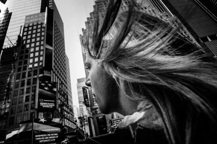 Black and white photo of a woman's windblown hair close to the lens with tall buildings behind her