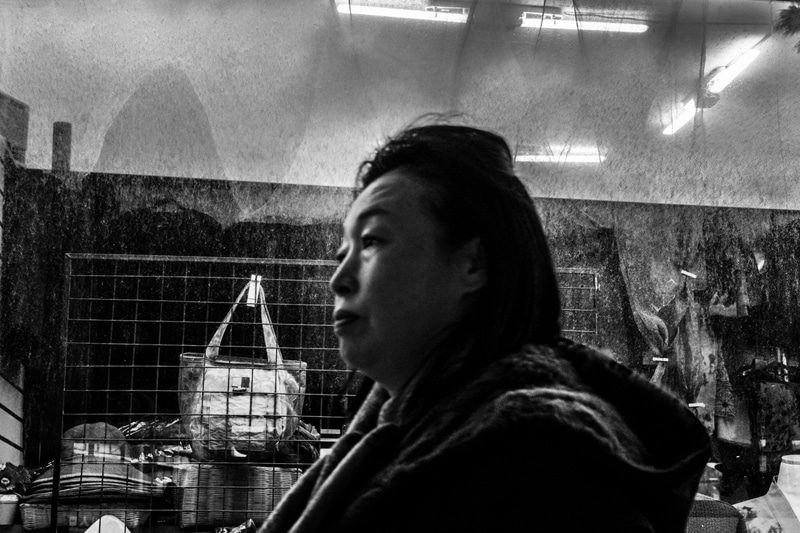 Black and white photo of a woman seen through a shop window, with lights and a handbag behind her