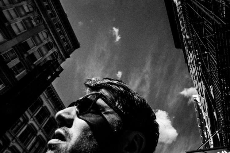 Low angle black and white photo of a man in sunglasses against tall buildings and sky