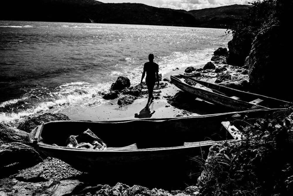 Black and white photo of a lone figure walking on a rocky beach behind wooden boats
