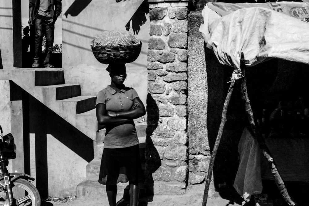 Black and white street photo of a woman standing with arms crossed by outdoor stairs as another woman carries a basket above