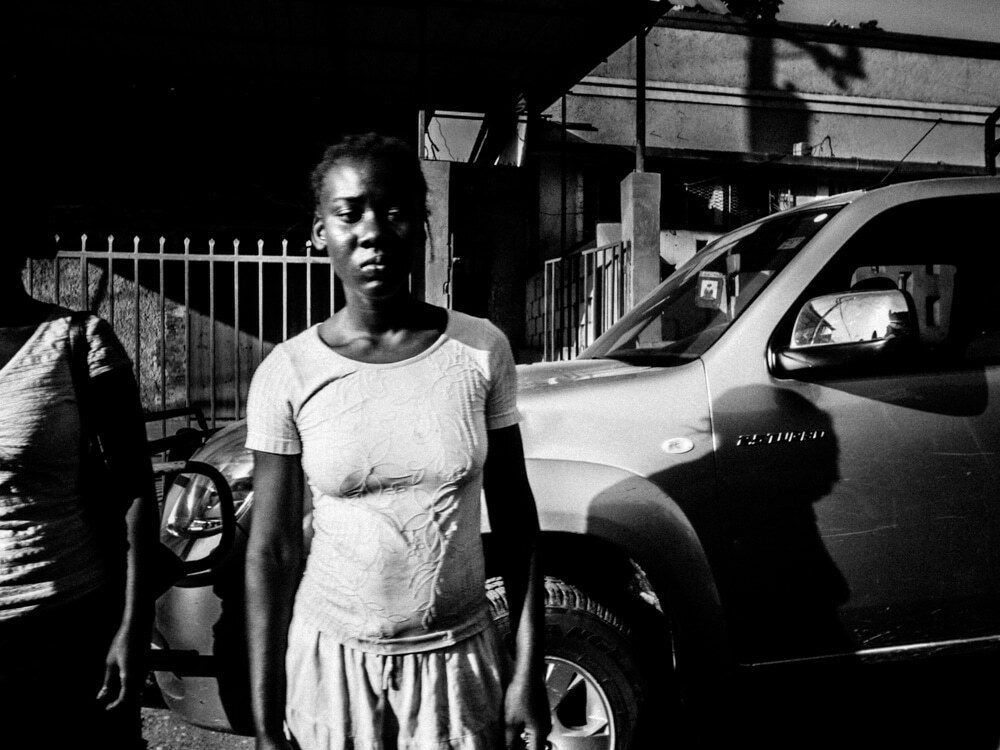 Woman in a t-shirt standing in a patch of hard sunlight next to a parked car, with a dark shaded background in Haiti