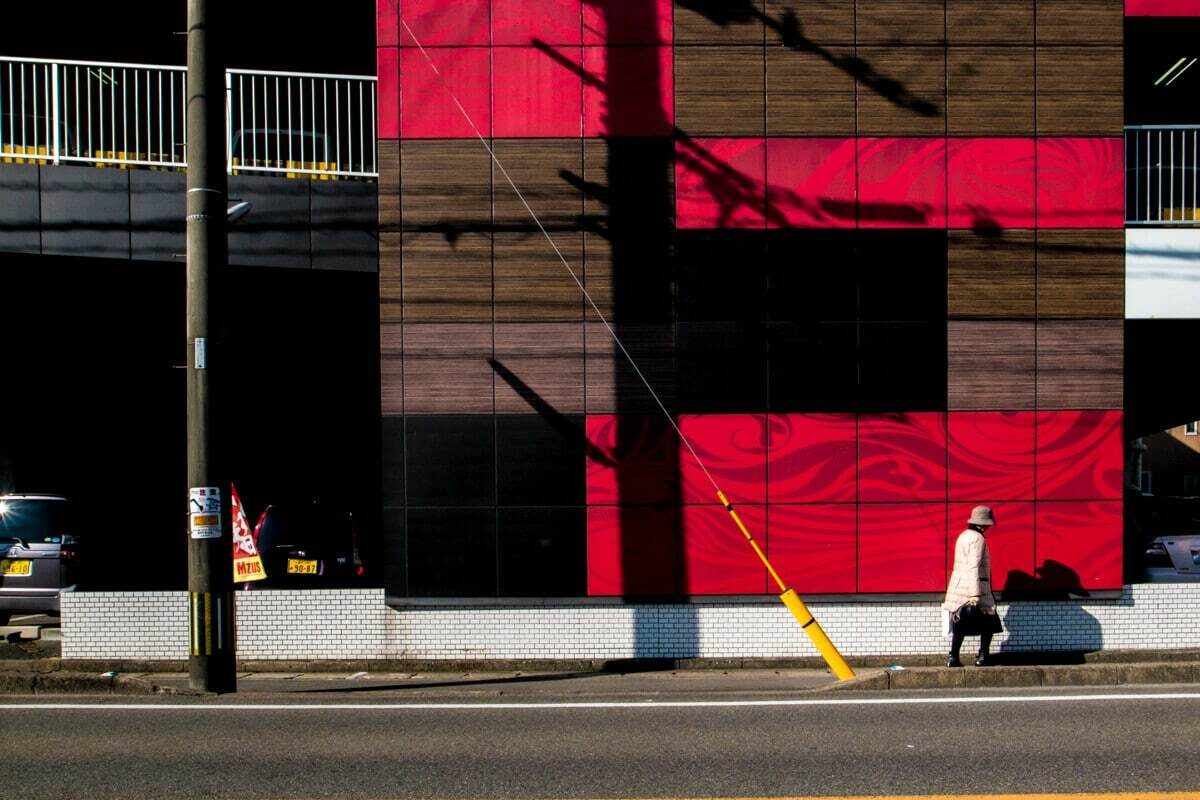 Colour street photo of a small figure walking past a large red and brown wall under a shadow of power lines