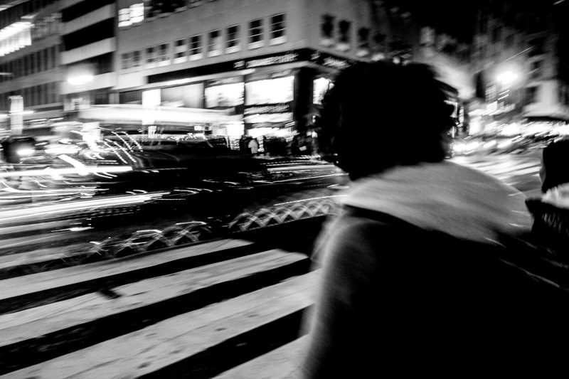 Black and white night photo of a person crossing a street as car lights streak behind