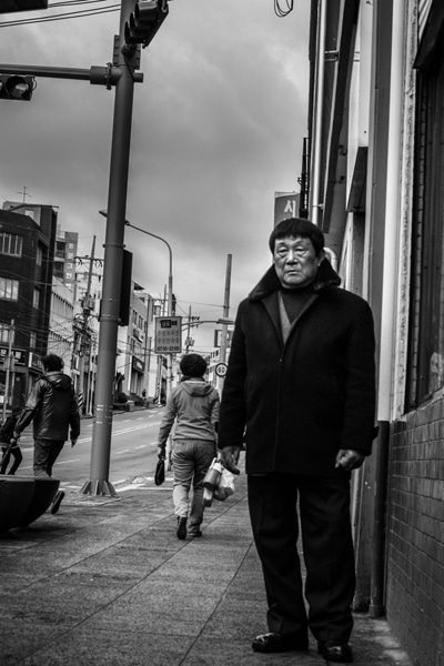 Vertical black and white photo of an older man in a dark coat standing on a city sidewalk
