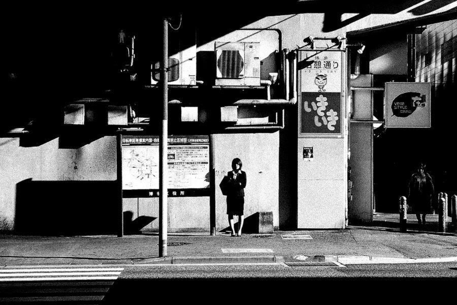High contrast black and white street photo of a small dark figure standing by a pole under a bright white building, with deep black shadow across the foreground