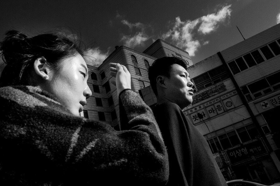 Black and white street photo of a young woman in the foreground and a man behind her, both looking to the side, with tall buildings and a bright sky behind them