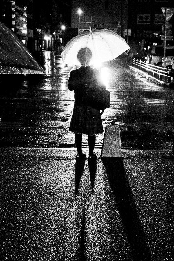 Black and white night street with a woman holding a glowing umbrella under a street lamp in the rain