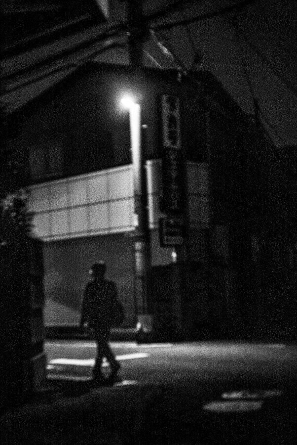Dark black and white night street with a lone figure walking through a small patch of light