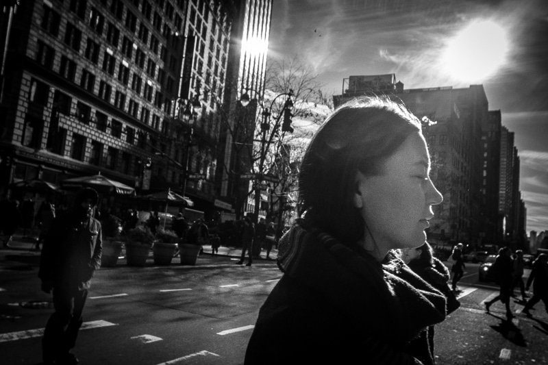 Black and white photo of a woman's face in hard sunlight in the foreground of a busy city street with tall buildings