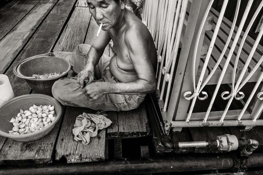 Black and white photo of an older woman smoking while peeling garlic on a wooden porch in Penang