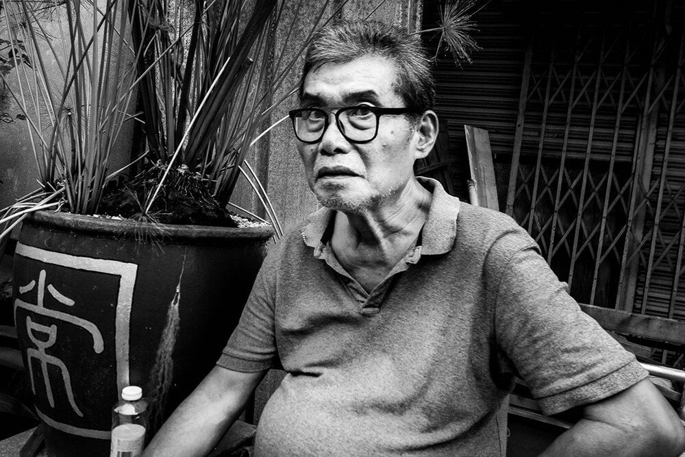 Black and white photo of an older man in glasses looking straight at the camera beside a large planter in Penang