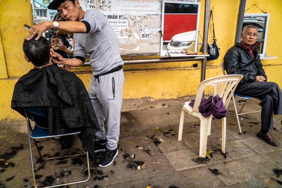 Colour candid photo of a street barber cutting a man's hair on a sidewalk against a yellow wall while another man sits and waits