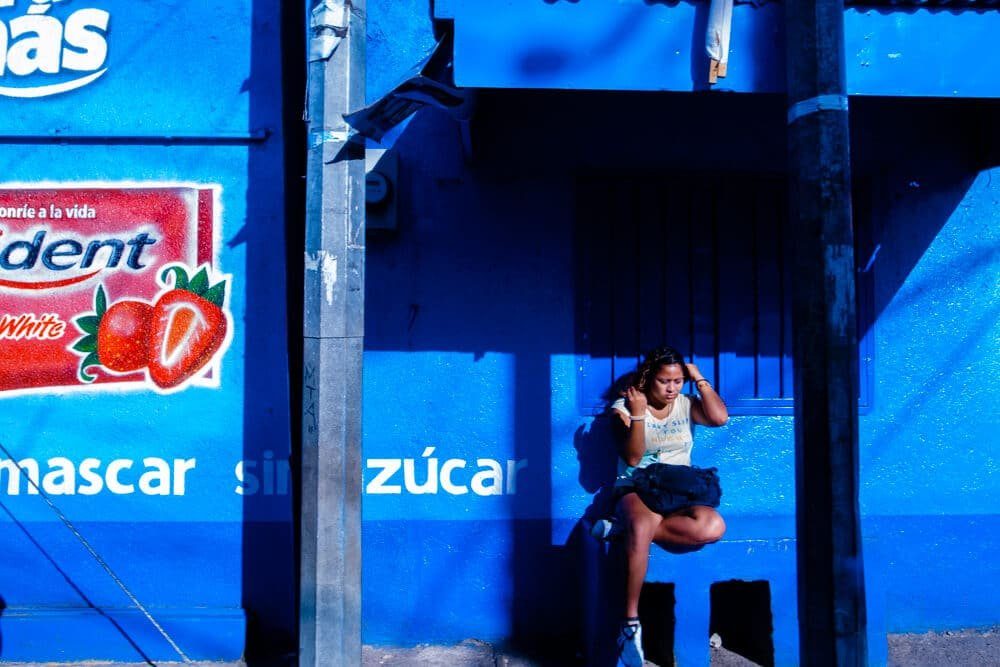 Street photo in Nicaragua of a woman sitting against a bright blue wall, with a red strawberry candy sign at the top left