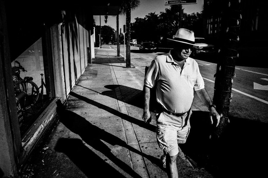 Black and white street photo of a man in a hat walking past a storefront, with hard diagonal shadows across the sidewalk