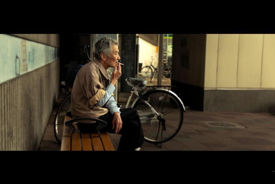 Cinematic color street photo of an old man smoking on a bench next to a bicycle, in warm muted tones