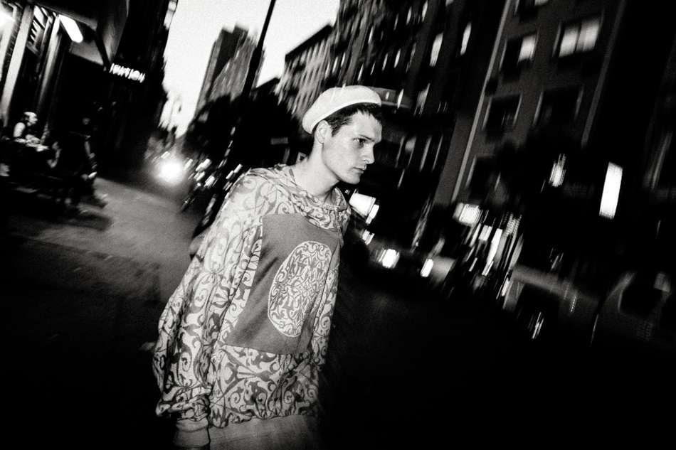 Young man in a patterned shirt and cap on a city street at night, blurred and tilted, with bright lit buildings behind