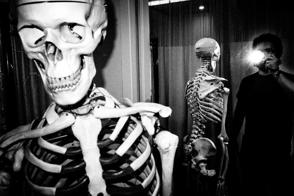 Anatomical skeleton model lit by hard flash in the foreground with a second skeleton and a man with a light in the background