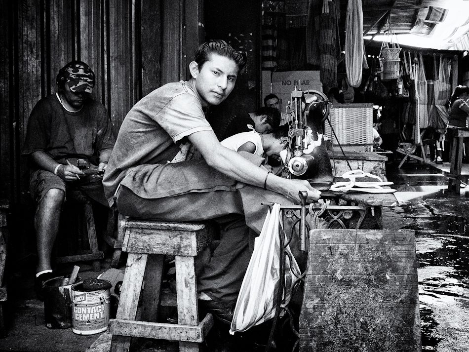 Black and white photo of a young man in a Nicaraguan market workshop lifting his head to look at the camera while working