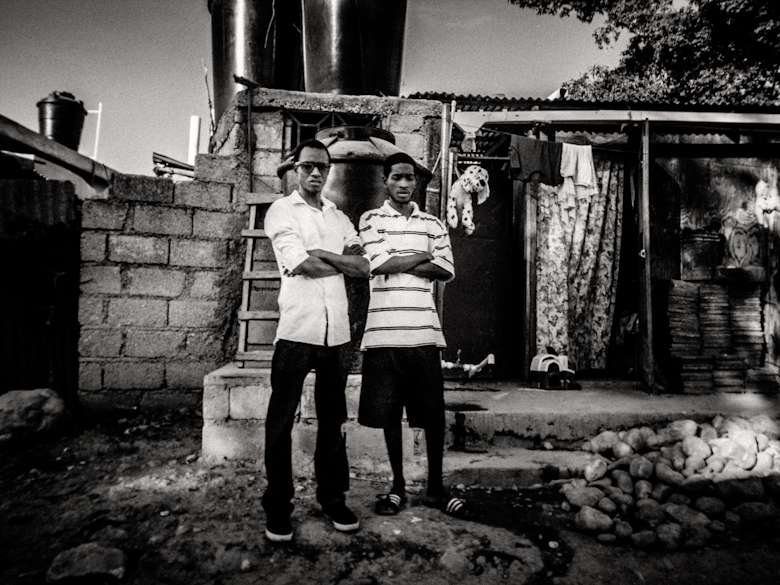 Two men standing in front of a corrugated metal shack, black and white