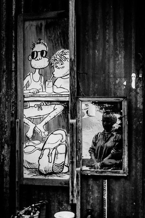 Black and white photo of a cartoon mural of two figures above a framed picture of a real man on a corrugated metal door in Penang
