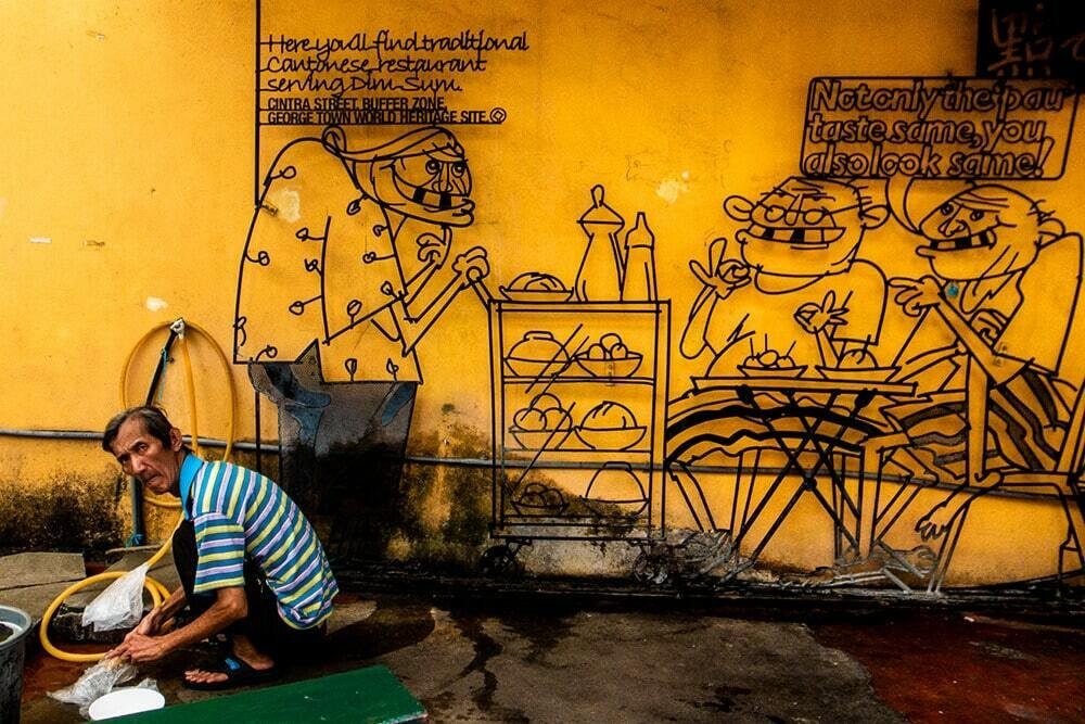 Man crouching to wash something under a large yellow mural of cartoon people eating at a table in Penang