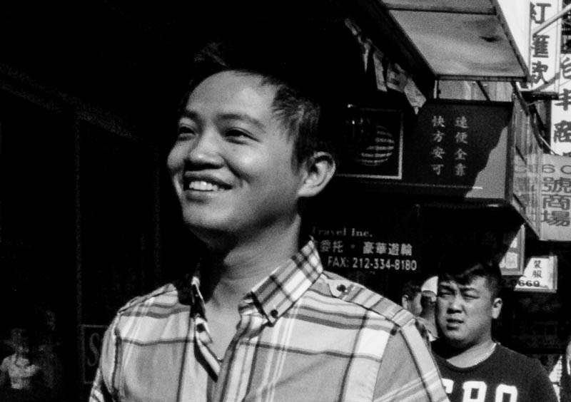 Smiling man in a checked shirt on a city street with shop signs behind him, black and white