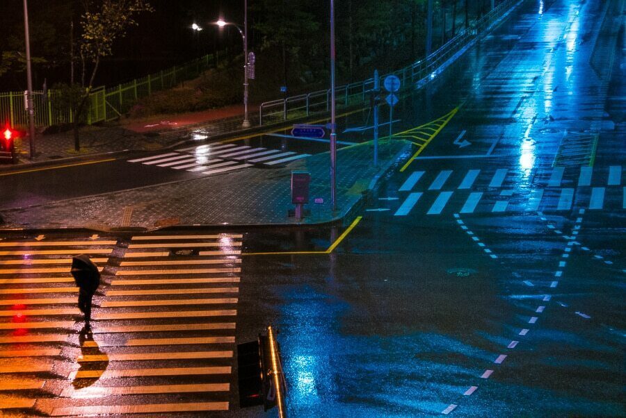 Seoul crosswalk at night with red and blue lights reflecting on the wet road and a pedestrian crossing