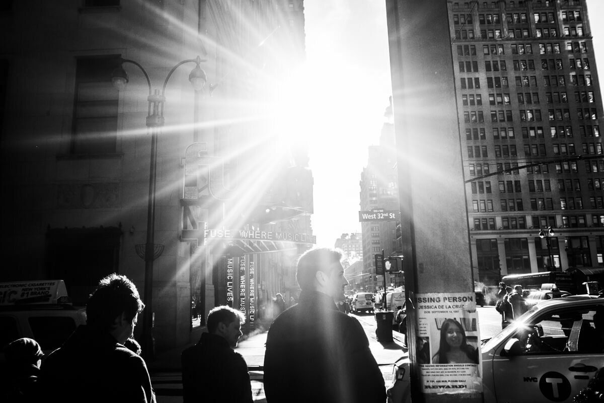 Black and white street scene shot into the sun, with a strong sunburst between buildings and people in silhouette