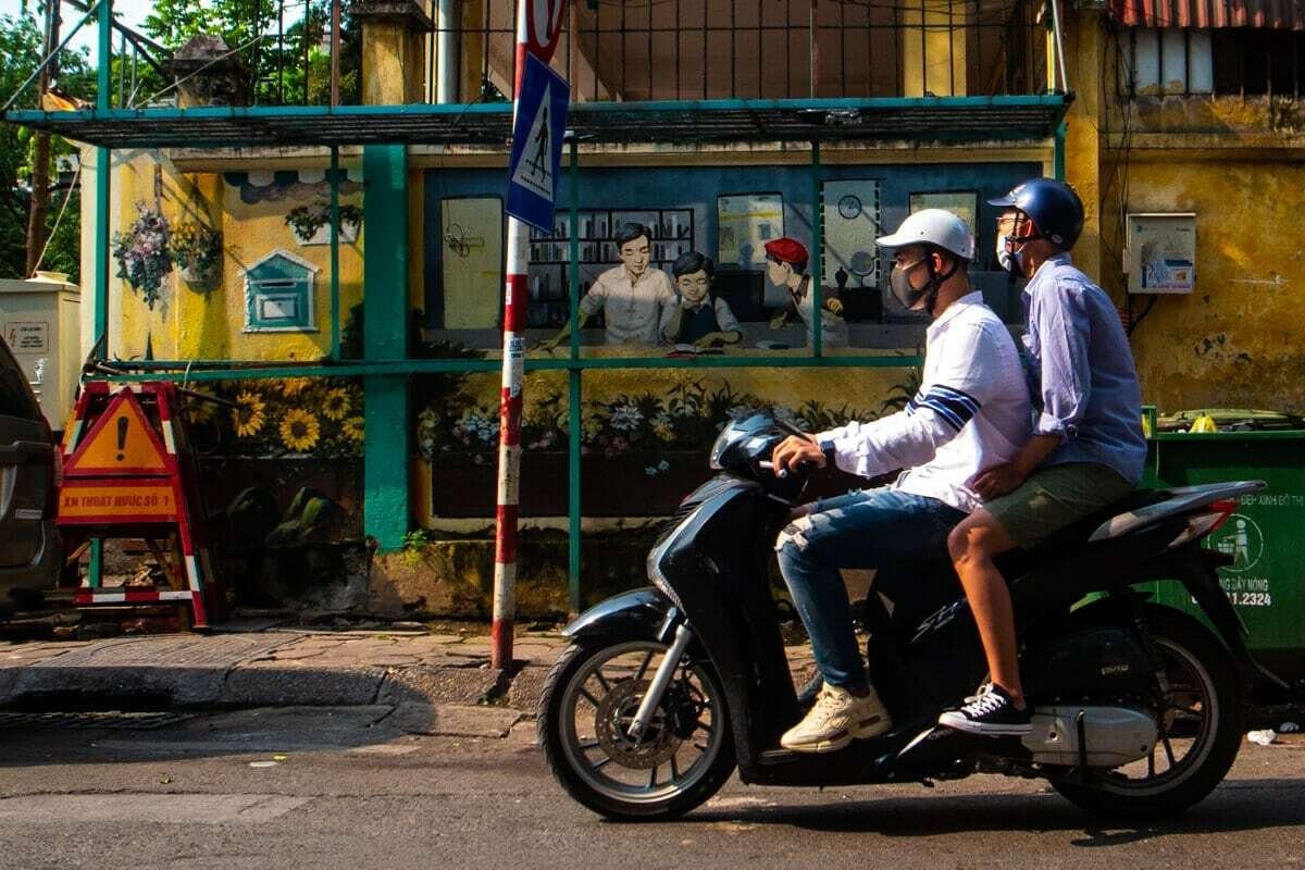 Two men in helmets ride a scooter past a colorful wall, sharp and frozen in motion