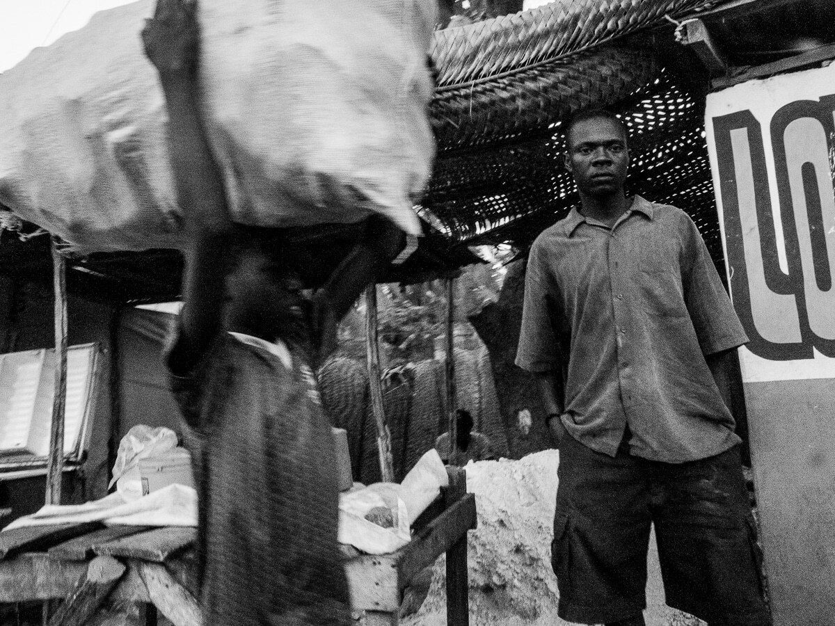 Black and white photo of a person carrying a huge sack on their head at a market, blurred by movement