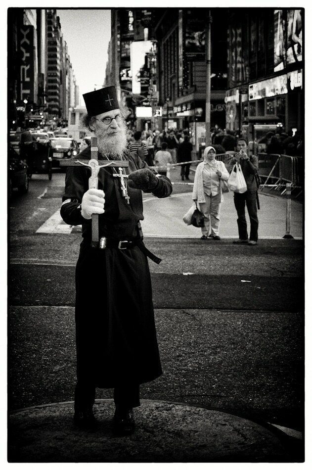Black and white street portrait of a bearded Orthodox priest holding a cross, standing in the middle of a busy New York street