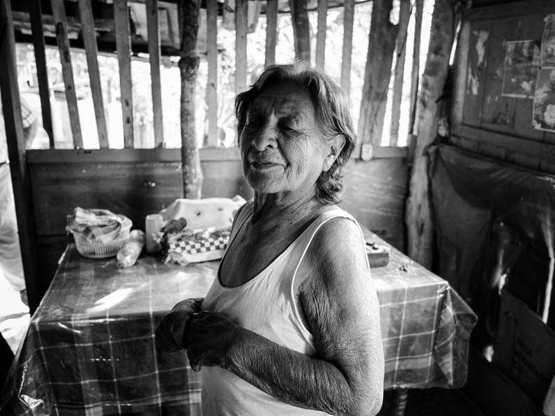 Black and white environmental portrait of an older woman in a tank top standing beside a table with a checked cloth in a wooden room