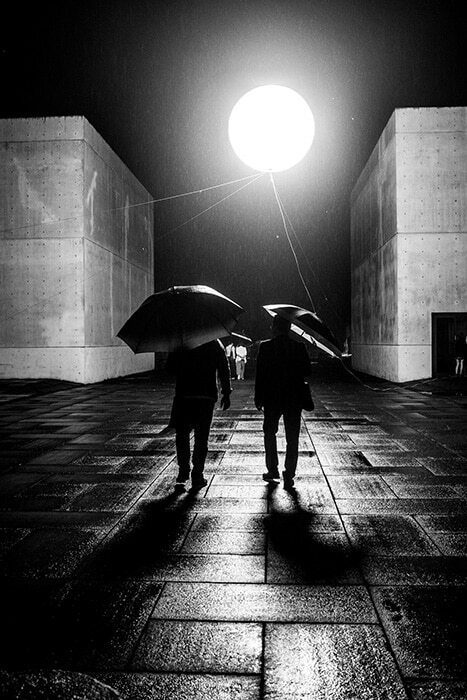 Black and white night photo of two people with umbrellas walking between two pale walls toward a large round light