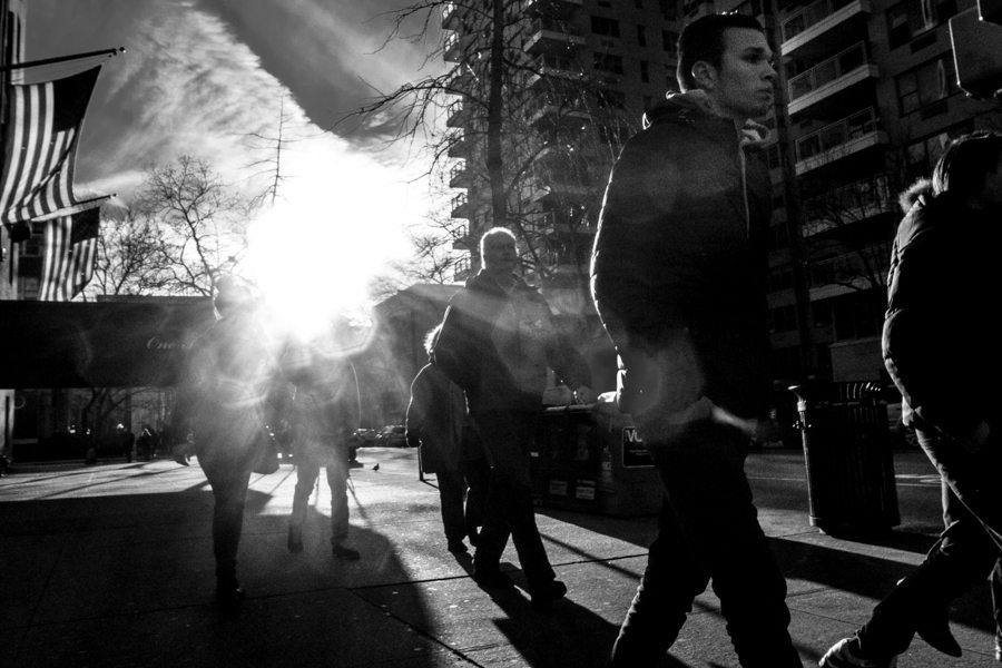 Black and white photo of the sun flaring between buildings behind pedestrians in silhouette, with an American flag on the left