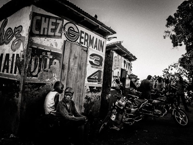 Grainy high-contrast black and white street scene in Haiti with men sitting outside a hand-painted wooden shop with motorbikes parked in front
