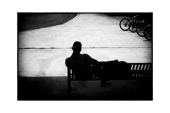 Black and white photo with black film-style borders of a man sitting on a bench in silhouette against a bright wall, bicycles at the edge of the frame