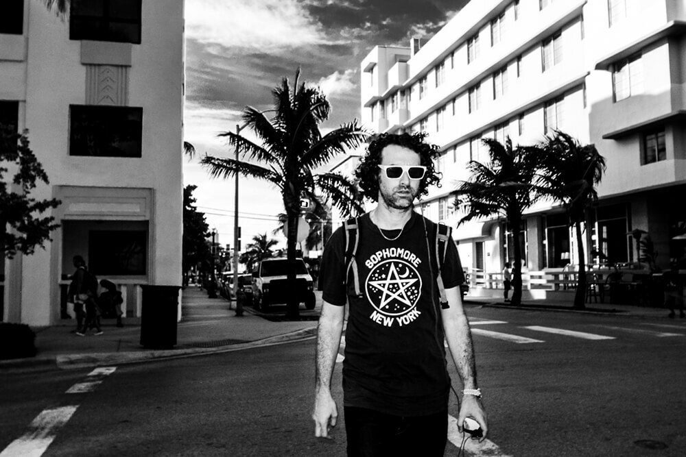 Black and white daylight flash photo of a man in sunglasses and a pentagram t-shirt crossing a Miami street